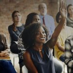 woman sitting in a classroom surrounded by classmates raising her hand to ask can anyone become a relationship coach